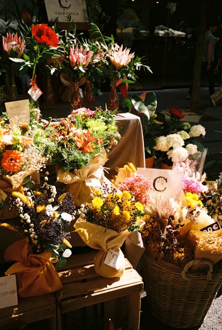 An assortment of fresh floral arrangements displayed atop wooden tables and in wicker baskets, featuring roses, gerbera daisies, protea, and various wildflowers in shades of pink, yellow, orange, red,