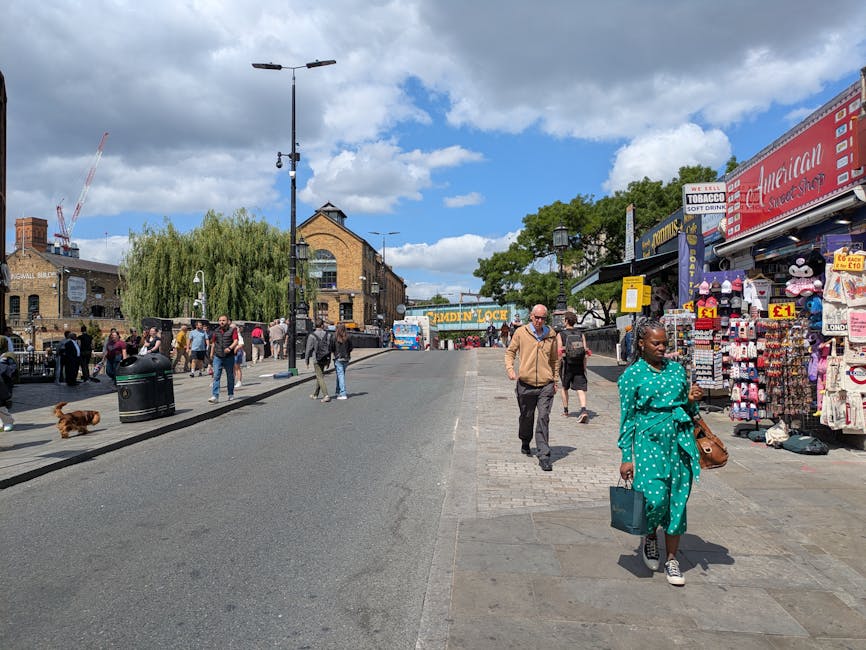 A lively street scene at Camden Market in NW1, featuring a variety of people strolling along the sidewalk. In the foreground, a woman dressed in a vibrant turquoise polka-dot outfit holds a brown hand