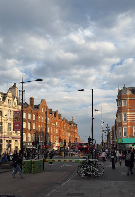 This image features a busy streetscape near Camden Town station, with historic brick buildings lining the road. The sky above is partly cloudy with patches of blue showing through. In the foreground, 