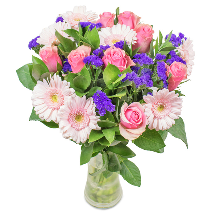 Displayed against a clean, bright background-a hallmark of contemporary Camden style-this floral arrangement features approximately seven to nine lush pink roses, some with gently curling edges just beginning to open, highlighting the distinctive spiral pattern typical of classic roses. Five to seven soft pink gerbera daisies, with their signature round faces and pale centers, add an airy brightness. Sprigs of intense, purple statice are woven among the blooms, bringing bold contrast and tactile variation-much like the dynamic street art of Camden's eclectic quarter. The foundation is built from abundant green foliage, with leaves both glossy and deep in tone, reminiscent of local parks and greenways such as Hampstead Heath. The flowers are presented in a clear glass vase, water line visible along the stems, contributing to a sense of freshness and immediacy. This arrangement, ready to brighten any Camden home, is an ideal gift for occasions from birthdays to Mother's Day, or simply as a colourful accent for a creative studio or office along Camden High Street. The feeling conveyed is one of lively celebration and affectionate connection, rooted in Camden's diverse, welcoming community.